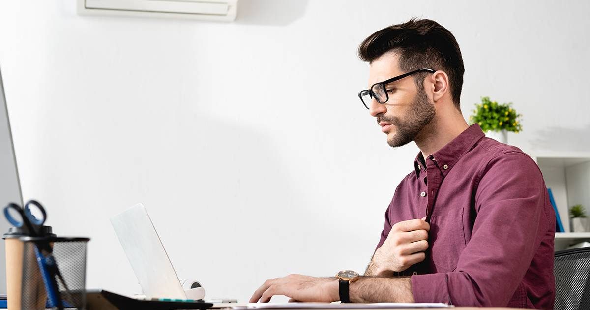 young businessman touching shirt while working on laptop and suffering from heat near air conditioner
